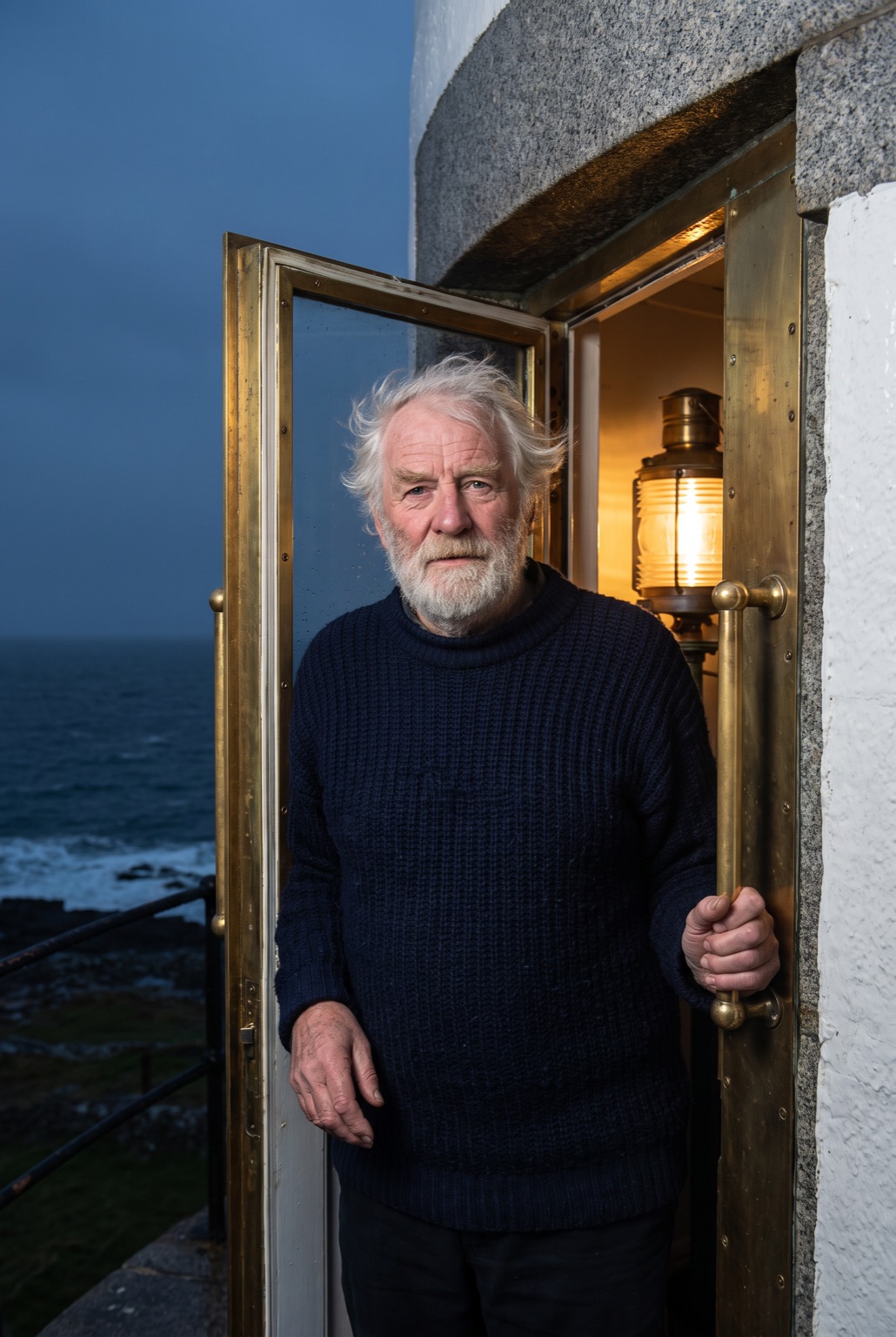 An older man with white hair and beard in a navy wool jumper, standing in the doorway of a lighthouse lantern room at night.