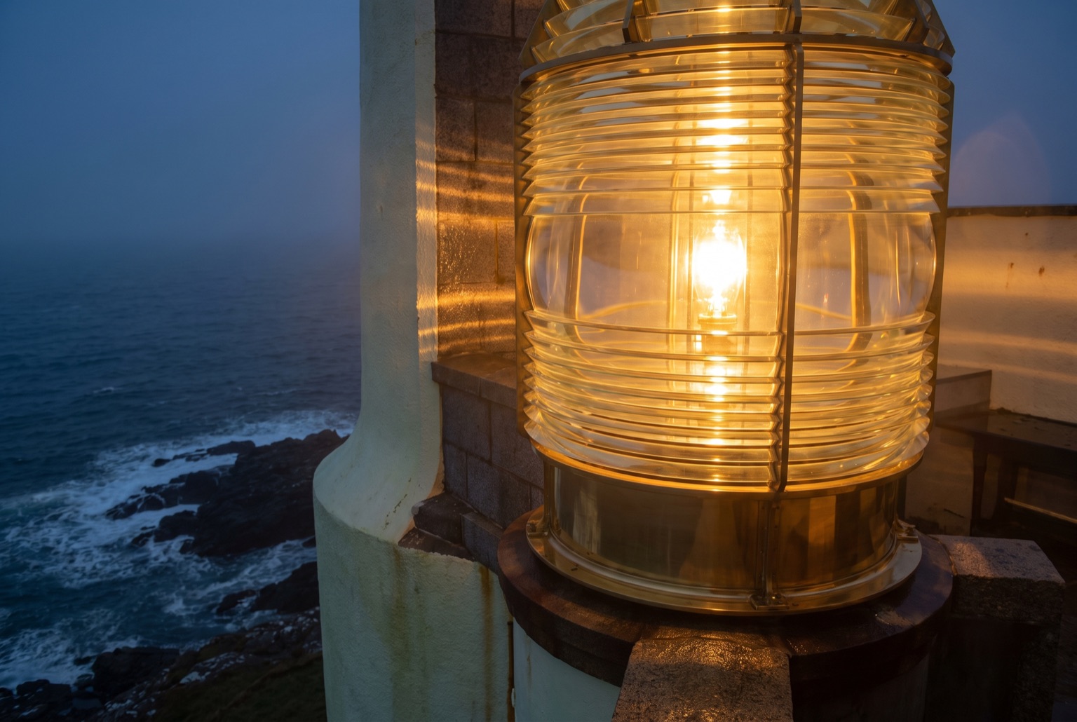 A large Victorian Fresnel lighthouse lens: concentric rings of ground glass prisms in a brass frame, lit warmly from within.