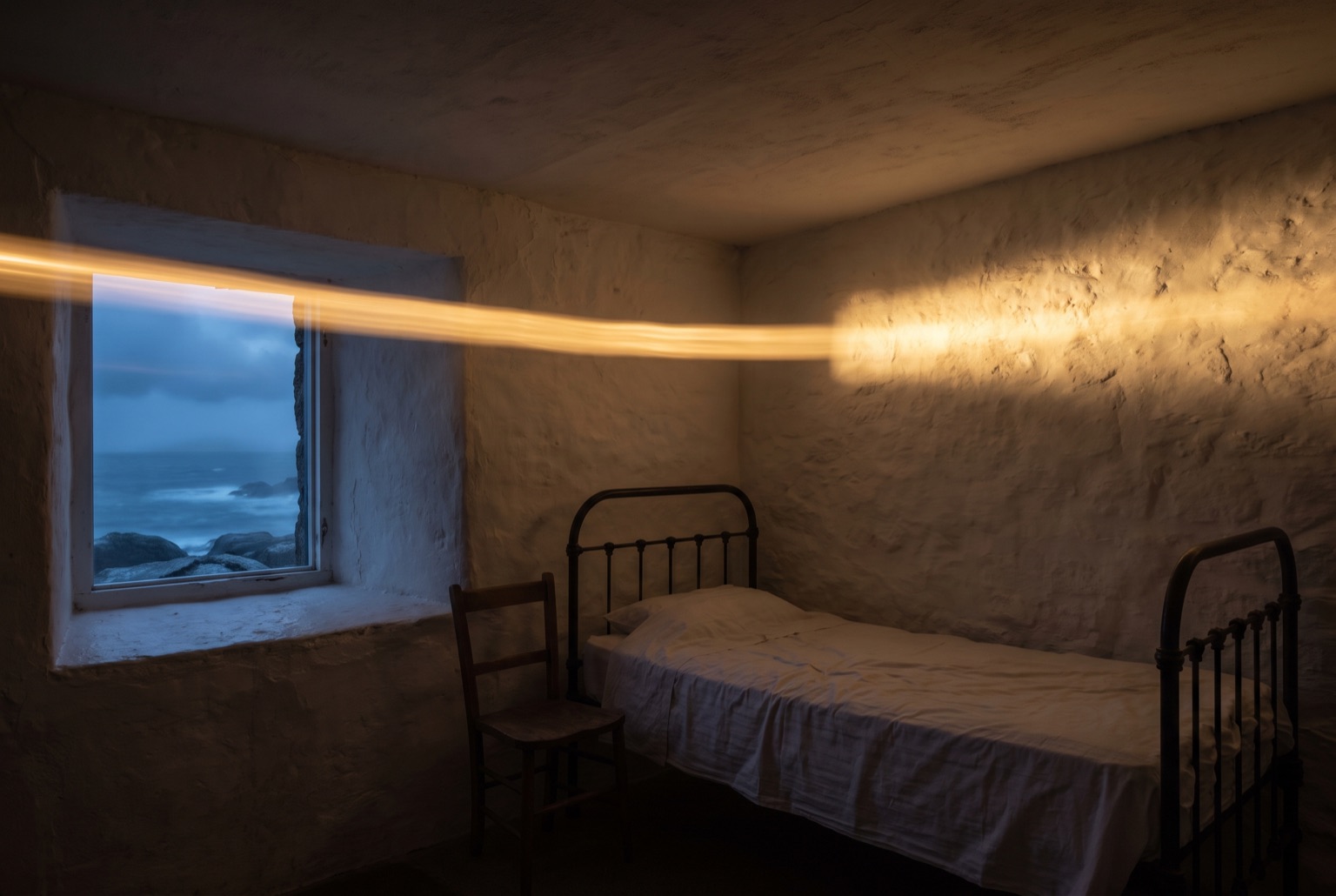 A whitewashed bedroom at night with an iron bedstead and a deep-set window, a band of warm light thrown across the ceiling by the lighthouse beam.
