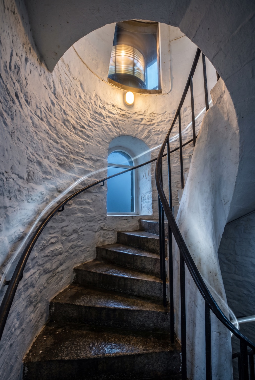 A spiral granite staircase inside a lighthouse tower, seen looking upward, with an iron handrail and a light at the top.