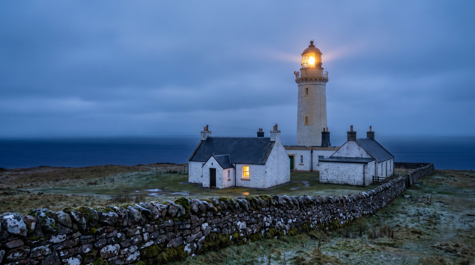 Low white cottages with slate roofs beside a pale lighthouse tower on open grassland at blue hour, one window lit, a drystone wall in front and the sea beyond.