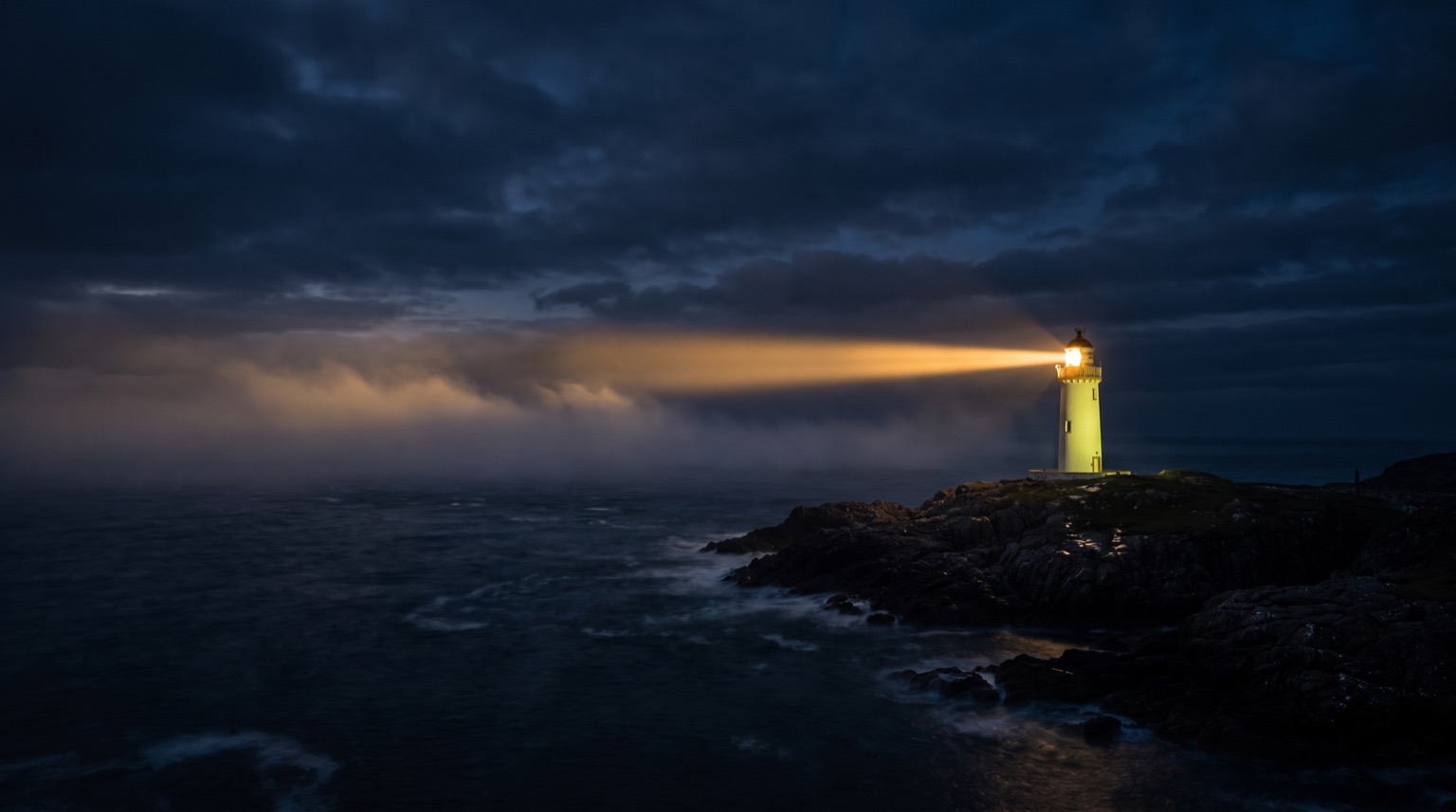 A lime-white lighthouse tower on a low headland at night, its warm beam cutting out over dark water into low cloud.
